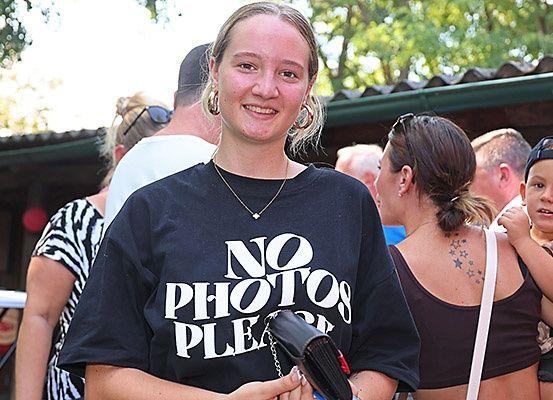 Eine Frau mit einem schwarzen T-Shirt, auf dem 'KEINE FOTOS BITTE' steht, lächelt und hält eine Geldbörse in einer überfüllten Gegend. Hinter ihr stehen mehrere Menschen, und eine Frau mit einem Tattoo auf dem Rücken trägt ein Kind.