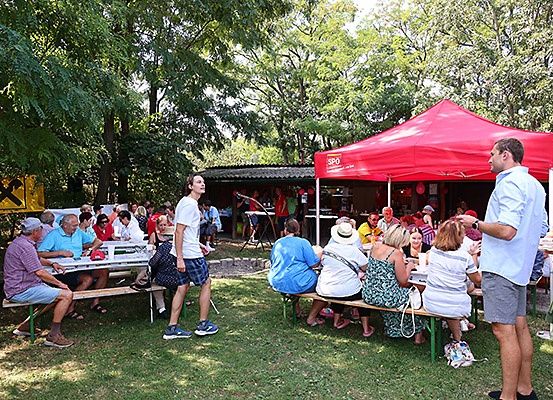 Eine Gruppe von Menschen sitzt an Picknicktischen in einem Park. Ein Mann steht neben einem roten Zelt. Bäume umgeben das Gelände, und ein Gebäude ist im Hintergrund zu sehen.