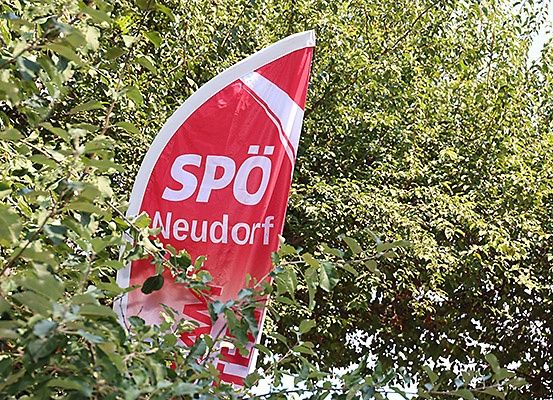 Ein rotes Banner mit weißem Text, das 'SPO Neudorf' steht, ist teilweise von grünem Laub verdeckt, mit hellem Sonnenlicht, das die Szene beleuchtet.