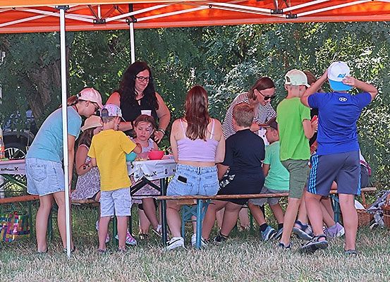 Eine Gruppe von Menschen, einschließlich Erwachsener und Kindern, versammelt sich unter einem orangefarbenen Zelt für ein Picknick. Einige sitzen an einem Tisch, während andere stehen, und ein Junge justiert seinen Hut.