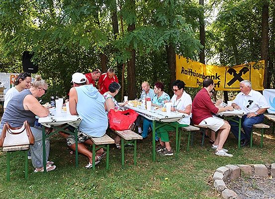 Eine Gruppe von Menschen sitzt an Tischen in einem Park und genießt ihre Mahlzeiten. Im Hintergrund befinden sich Bäume und ein Banner.