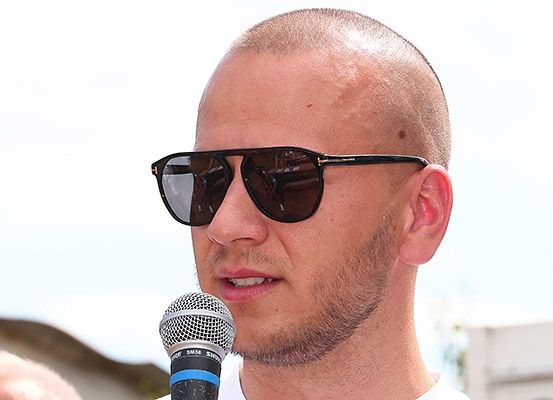 Ein Mann ist im Freien, trägt eine Sonnenbrille und spricht in ein Mikrofon. Er ist kahl und hat einen Bart.