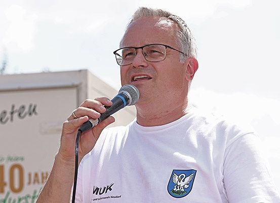 Ein Mann in einem weißen T-Shirt mit einem Logo auf der linken Brustseite hält ein Mikrofon und spricht. Hinter ihm zeigt ein Gebäude die große Zahl 40.