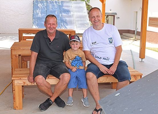 Drei Männer sitzen auf einer Holzbank im Freien, einer trägt eine Kappe und die anderen lächeln. Die Umgebung scheint ein Hinterhof zu sein.