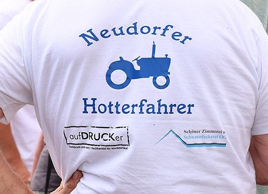Ein weisses T-Shirt mit einem blauen Traktor-Bild und den Worten Neudorfer Hotterfahrer, gedruckt von aufDRUCKer. Unter dem Traktor befindet sich ein Emblem mit dem Text Schöner Zimmerei u. Schwarzdeckerei OG.