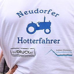 Ein weisses T-Shirt mit einem blauen Traktor-Bild und den Worten Neudorfer Hotterfahrer, gedruckt von aufDRUCKer. Unter dem Traktor befindet sich ein Emblem mit dem Text Schöner Zimmerei u. Schwarzdeckerei OG.