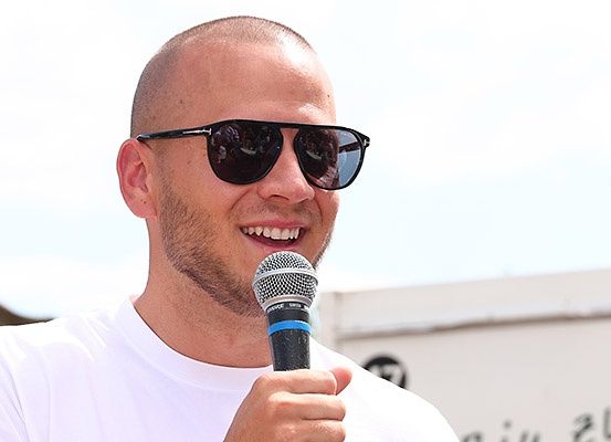 Ein Mann mit kahlgeschorenem Kopf, der eine Sonnenbrille und ein weißes T-Shirt trägt, lächelt und ein Mikrofon in der Hand hält.