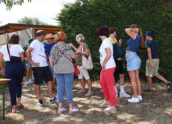 Eine Gruppe von Menschen in Freizeitkleidung steht in einem Außenbereich, wahrscheinlich bei einer Veranstaltung, mit einem Holztisch und einer Hecke im Hintergrund.