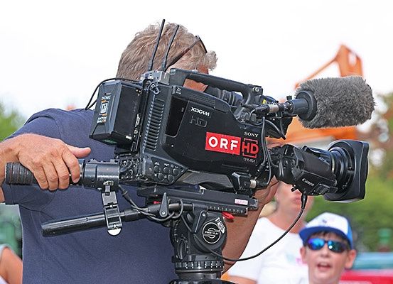 Ein Mann mit einer Sony HD-Kamera mit ORF HD-Logo filmt, wobei ein Mikrofon angeschlossen ist, während andere im Hintergrund zusehen.
