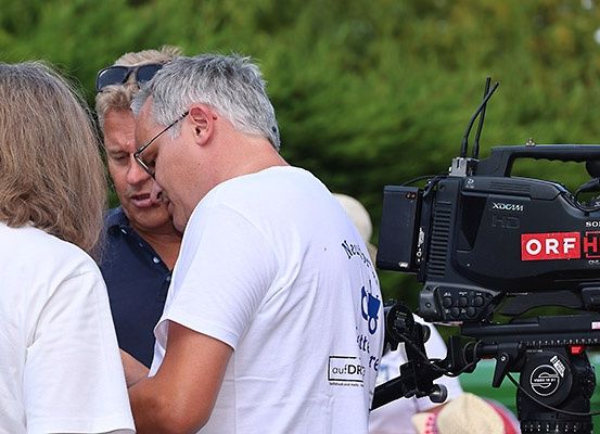 Drei Menschen sind draußen, einer von ihnen filmt mit einer Kamera. Der Mann in Weiß trägt eine Brille und schaut etwas an. Hinter ihm schaut eine Frau zur Kamera.