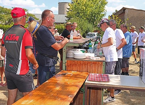 Eine Gruppe von Männern steht um einen Essensstand herum, einige tragen Hüte, Brillen und Sneakers. Ein Mann in einem roten Shirt mit dem Text 'KTM' steht links.