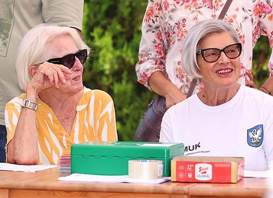 Zwei ältere Frauen sitzen an einem Tisch mit einer grünen Box und einer roten Box mit der Aufschrift 'Silol'. Sie lächeln, eine trägt eine Brille und einen Ring. Hinter ihnen steht eine Frau mit einem Blumenshirt und einer braunen Tasche.