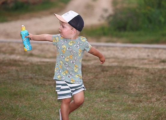 Ein junger Junge in einem gestreiften Hut und Shirt spielt draußen, eine blaue Flasche in der Hand haltend und nach links schauend.