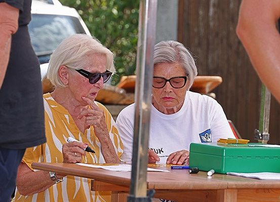 Zwei ältere Frauen sitzen an einem Tisch und schreiben. Eine trägt eine Sonnenbrille und ein gelbes Shirt, die andere ein weißes Shirt. Auf dem Tisch liegen eine grüne Box, Stifte und Papiere. Eine Person steht hinter ihnen.