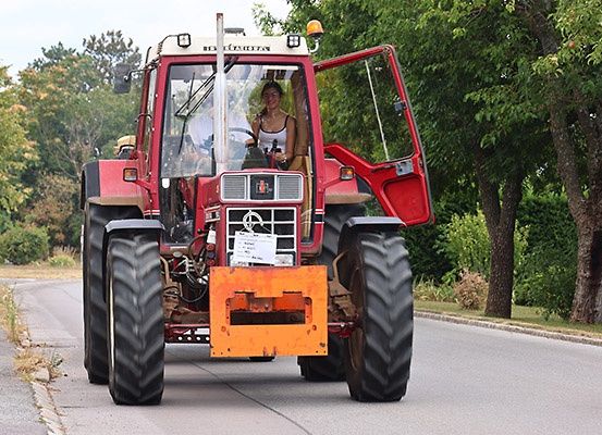 Eine Frau fährt einen roten Traktor auf einer asphaltierten Straße, mit Bäumen und Grünpflanzen auf beiden Seiten.