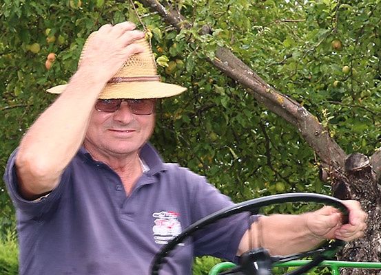 Ein älterer Mann mit Strohhut und Sonnenbrille fährt in einem Obstgarten. Er hält das Lenkrad mit beiden Händen.