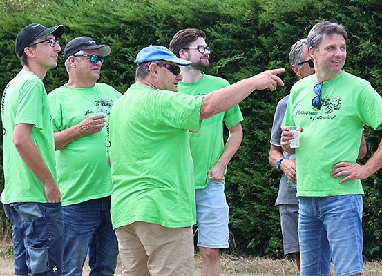 Fünf Männer in grünen T-Shirts, einer mit einem Getränk, stehen vor einer Hecke, wobei einer der Männer mit Gesten zeigt und deutet.