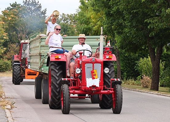 Eine Gruppe von Menschen fährt auf einem roten Traktor mit Anhänger auf einer von Bäumen gesäumten Straße.
