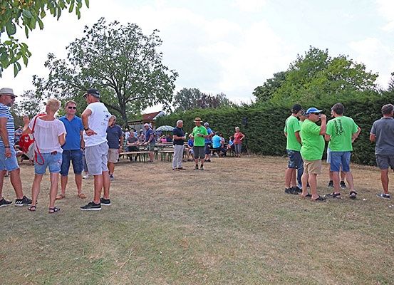 Mehrere Menschen stehen in einem Grasbereich, einige in grünen Shirts. Hinter ihnen sind Tische und Stühle unter Schirmen aufgestellt. Bäume und Büsche sind im Hintergrund.
