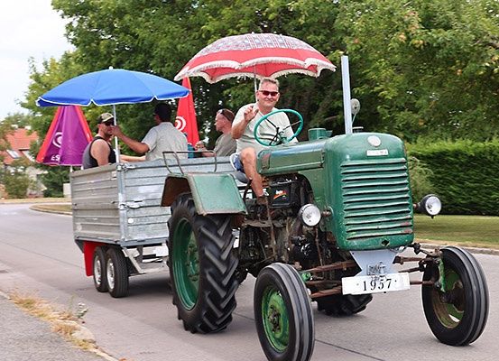 Ein grüner Oldtimer-Traktor zieht einen Anhänger mit vier Personen unter Schirmen. Ein Mann fährt und drei sitzen im Anhänger.