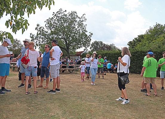 Menschen versammeln sich an einem sonnigen Tag im Freien, einige in Freizeitkleidung und Hüten, mit Tischen und Bänken im Hintergrund, Bäume und Büsche umgeben den Raum.