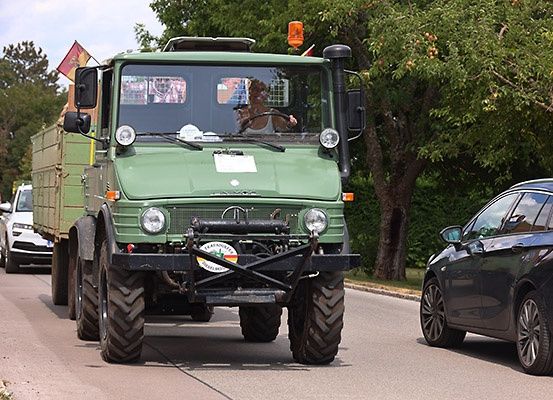 Ein grüner Militärlastwagen fährt mit einer Flagge auf dem Dach und einem Aufkleber am Stoßfänger auf einer Straße.