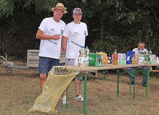 Zwei Männer stehen hinter einem Tisch mit Getränken darauf. Sie tragen weiße T-Shirts mit Text auf der Vorderseite. Ein anderer Mann sitzt am Tisch. Bäume sind im Hintergrund.
