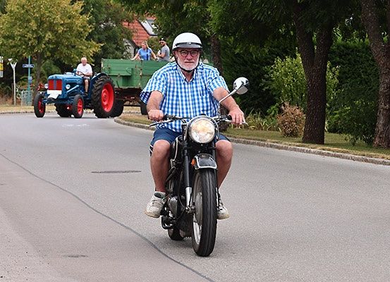 Ein Mann auf einem Motorrad fährt die Straße hinunter, während ein blauer Traktor mit Menschen hinten drinnen hinter ihm fährt.