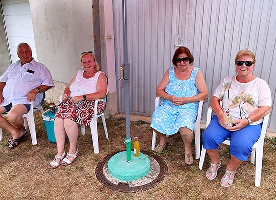 Vier ältere Menschen sitzen in Stühlen um eine runde Basis. Drei Frauen tragen Sonnenbrillen, während ein Mann eine Uhr trägt. Eine Frau hält eine Flasche, und ein Eimer steht in der Nähe.