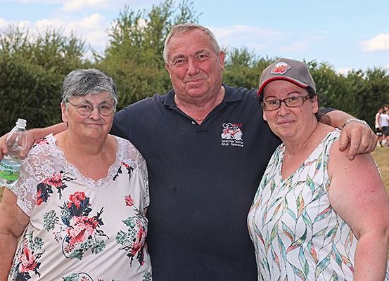 Drei Menschen stehen zusammen in einem Außenbereich. Der Mann hat seine Arme um die beiden Frauen gelegt. Eine Frau hält eine Flasche. Hinter ihnen stehen mehrere Menschen, und einige Bäume befinden sich in der Umgebung.