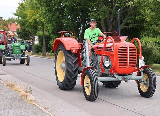 Ein junger Mann fährt einen roten Traktor die Straße hinunter, hinter ihm fahren zwei grüne Traktoren.