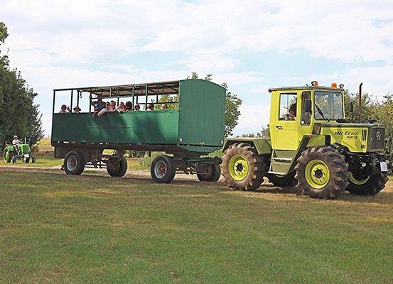 Ein gelber Traktor zieht einen grünen Anhänger mit Menschen im Inneren. Ein weiterer Traktor ist auf der linken Seite geparkt. Bäume und Büsche sind im Hintergrund zu sehen.