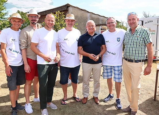 Eine Gruppe von sechs Männern, alle in verschiedenen Arten von Freizeitkleidung gekleidet, lächeln für ein Foto. Ein Mann in der Mitte hält ein Glas. Hinter ihnen steht ein Lkw und einige Bäume.