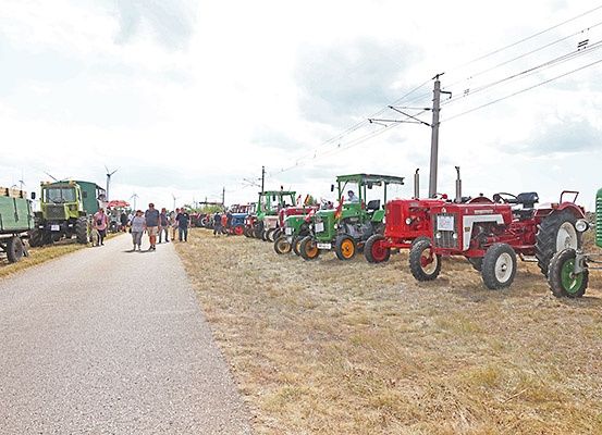 Eine Reihe von Oldtimer-Traktoren auf einem Grasfeld geparkt. Menschen gehen auf der Straße neben den Traktoren. Elektrische Masten und Drähte im Hintergrund.