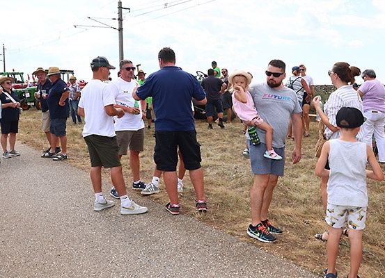 Eine Gruppe von Menschen steht am Straßenrand, einige tragen Hüte und Sonnenbrillen. Ein Mann hält ein Kind auf seiner Schulter. In der Nähe sind Maschinenfahrzeuge geparkt.