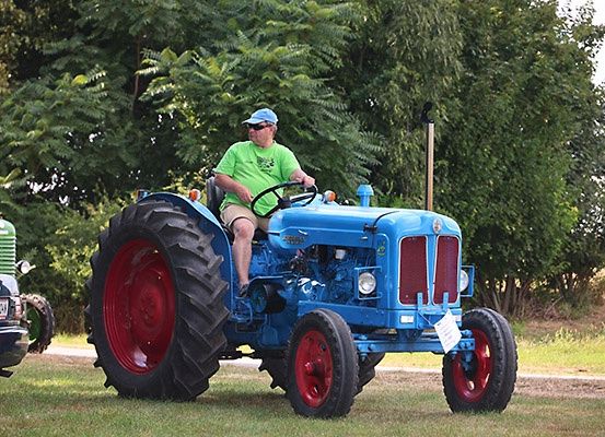 Ein Mann fährt einen blauen Traktor auf einem Feld mit grünen Bäumen im Hintergrund.
