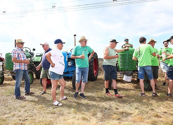 Eine Gruppe von Männern in grünen T-Shirts, Hüten und Sandalen steht vor Traktoren auf einem Feld, unterhält sich und trinkt Getränke.