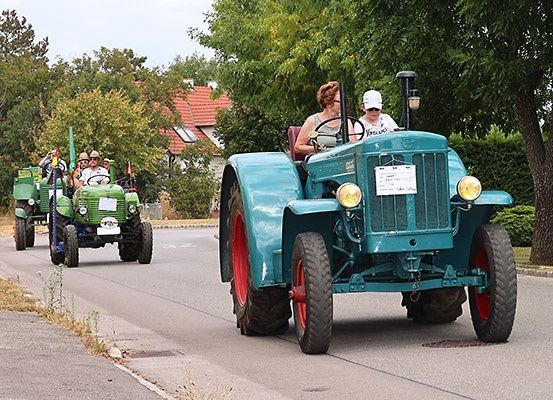 Eine Gruppe von Menschen fährt mit Oldtimer-Traktoren auf einer Straße, umgeben von Grün und einem Haus mit rotem Dach.