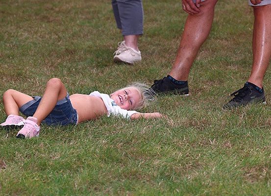 Ein kleines Mädchen liegt lächelnd auf dem Gras, mit ausgestreckten Armen. Zwei Erwachsene stehen hinter ihr, einer trägt Turnschuhe.