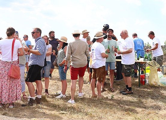 Eine Gruppe von Menschen in Freizeitkleidung versammelt sich auf einer grasbewachsenen Fläche, unterhält sich und hält Getränke in den Händen. Ein Tisch mit Flaschen ist in der Nähe aufgestellt. Der Himmel ist teilweise bewölkt.