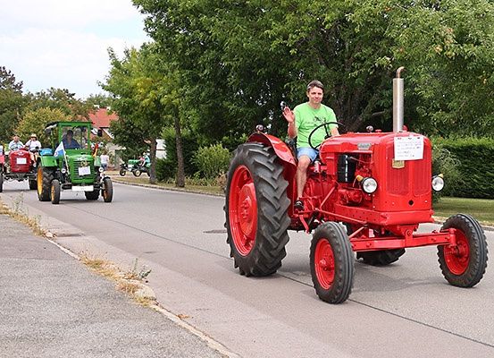 Ein Mann fährt einen roten Traktor auf einer Straße, mit anderen Traktoren und Menschen dahinter. Bäume und Büsche säumen die Straßenseiten.