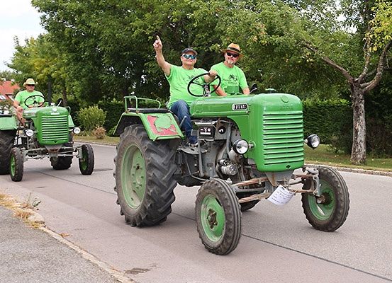 Drei Männer in grünen T-Shirts fahren auf grünen Traktoren eine Straße hinunter. Ein Mann zeigt. Bäume und Büsche säumen die Straße.