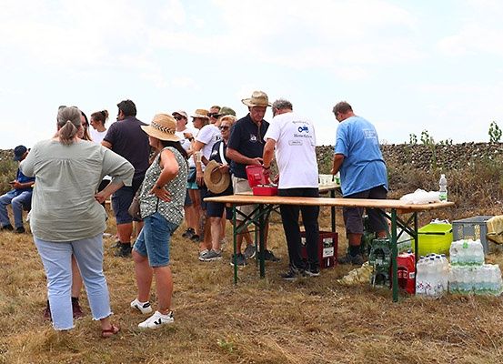 Eine Gruppe von Menschen versammelt sich auf einem Feld mit einem Tisch und Bänken, einige tragen Hüte und Sonnenbrillen. Eine Person beugt sich über den Tisch.