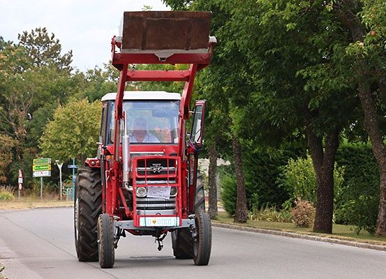 Ein roter Traktor mit einem großen Anbaugerät fährt die Straße hinunter, mit zwei Personen im Inneren. Bäume und ein Gebäude stehen am Straßenrand.