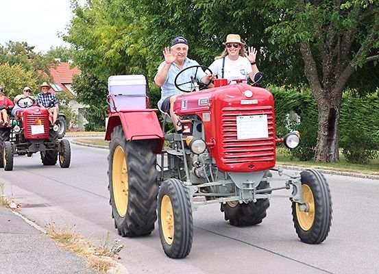 Eine Gruppe von Menschen in roten Traktoren fährt eine Straße entlang. Zwei Traktoren sind vorne, einer etwas dahinter. Bäume und Büsche säumen die Seiten.