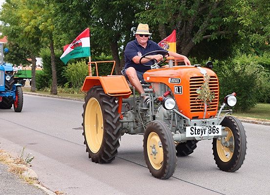 Ein Mann fährt einen orangefarbenen Oldtimer-Traktor mit ungarischen Flaggen auf der Straße. Hinter ihm ist ein blauer Traktor. Bäume und Häuser befinden sich an den Seiten.
