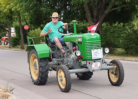 Ein Mann in einem grünen T-Shirt fährt einen Oldtimer-Traktor auf einer Straße mit Bäumen und einer Flagge an einem sonnigen Tag.
