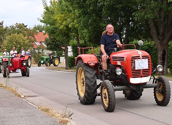 Eine Gruppe von Menschen fährt mit alten roten Traktoren auf einer Straße mit Bäumen auf beiden Seiten.
