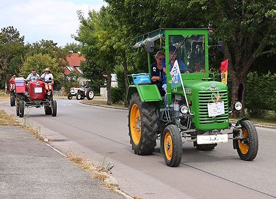 Mehrere Oldtimer-Traktoren fahren eine Landstraße hinunter. Ein grüner Traktor führt mit einer Flagge, gefolgt von einem roten Traktor. Bäume säumen die Straße, und Häuser sind in der Ferne sichtbar.
