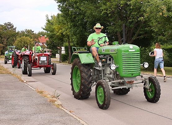 Eine Reihe von Oldtimer-Traktoren fährt eine Landstraße hinunter, eine Frau geht in der Nähe. Bäume und Häuser sind im Hintergrund.
